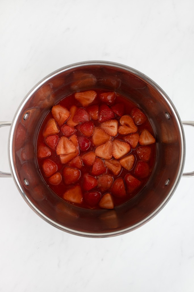 A stainless steel pot containing thickened strawberry mixture on a white marble surface.
