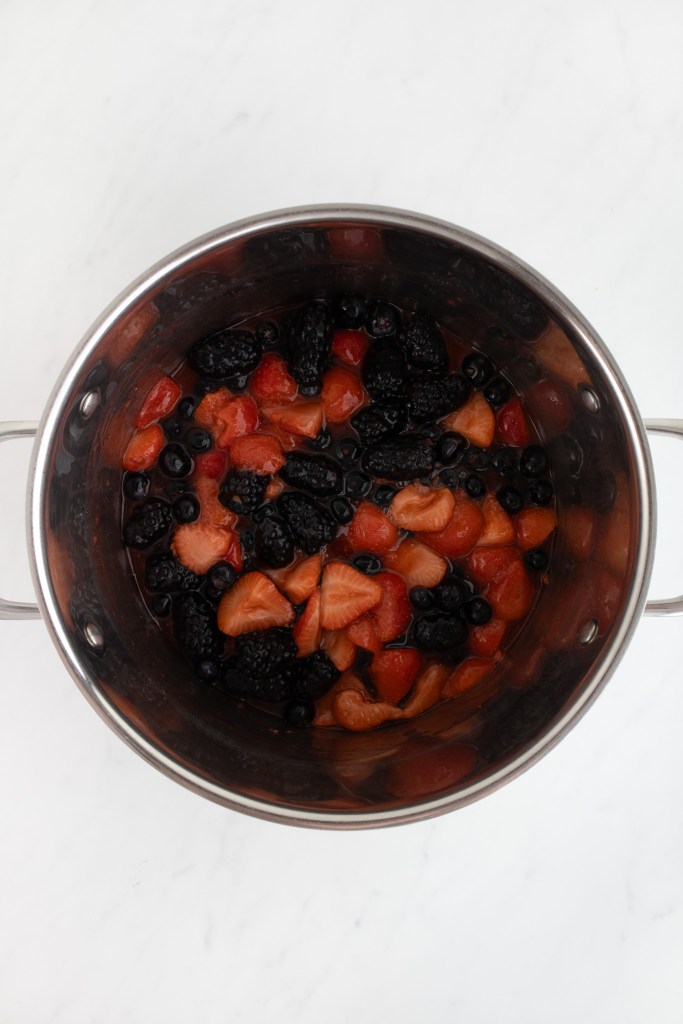 A stainless steel pot containing strawberry mixture mixed with blackberries and blueberries on a white marble surface.