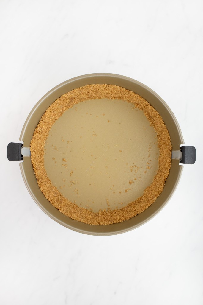 A springform pan with an even layer of graham cracker crust pressed around the edges, on a white marble surface.