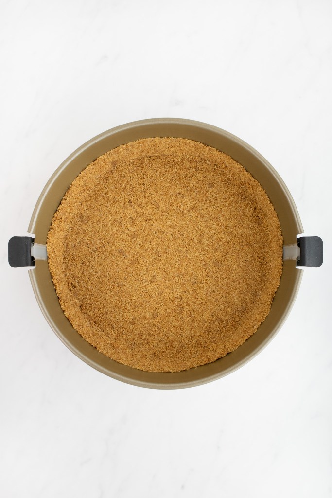 A springform pan with a pressed graham cracker crust sits on a white marble surface.
