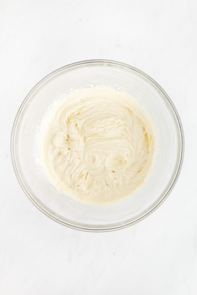A glass bowl filled with smooth cream cheese filling on a white marble surface.