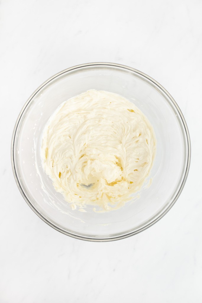 A glass bowl containing beaten cream cheese sits on a white marble surface, viewed from above.