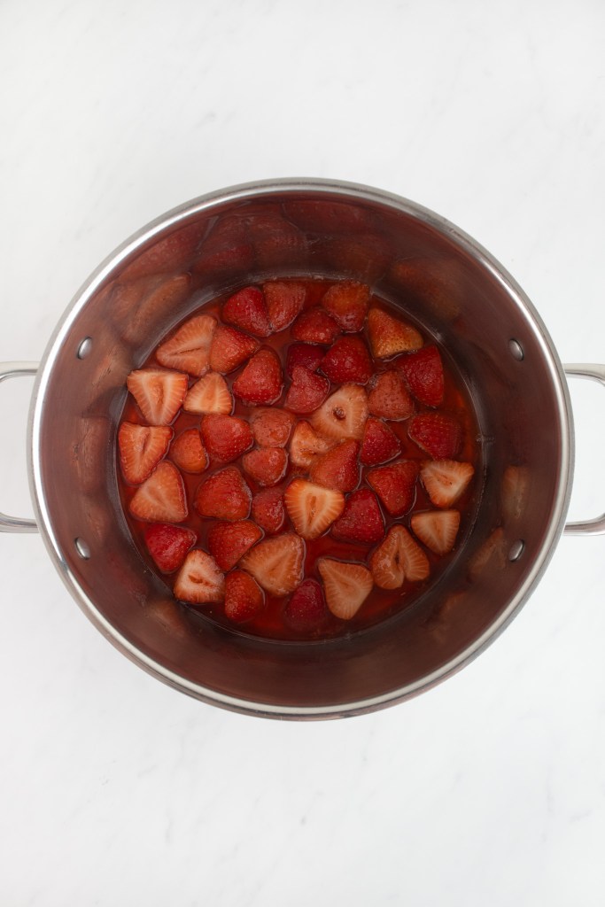 A stainless steel pot containing chopped strawberries in liquid sits on a white marble surface.