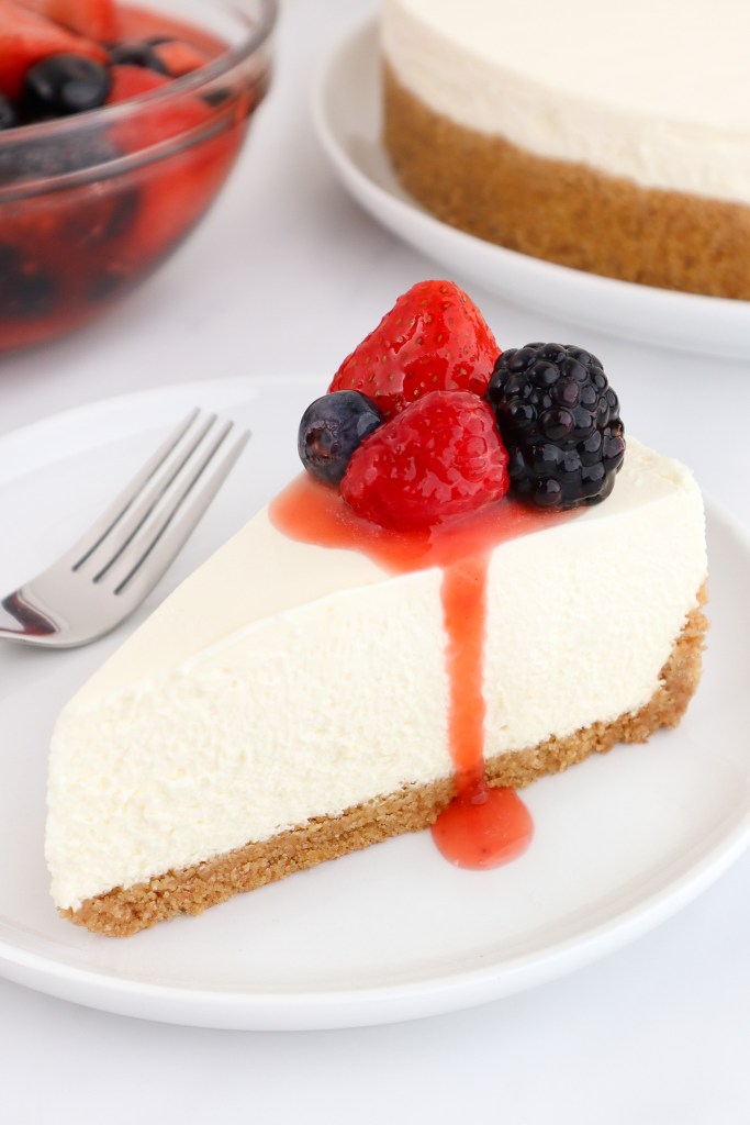 A slice of no bake cheesecake with mixed berry topping on top sits on a white plate with more cheesecake and a bowl of berry topping in the background