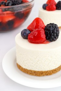 A small round no bake cheesecake with mixed berry topping on top sits on a white plate with a second mini cheesecake and a bowl of berry topping in the background.