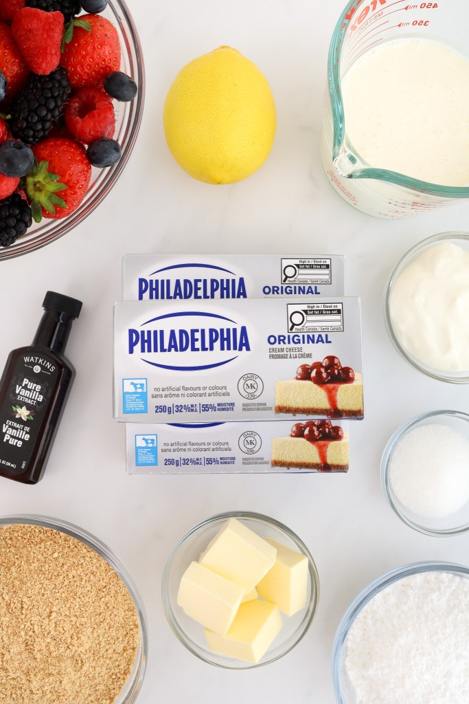 The ingredients for making no bake cheesecake including: mixed berries, lemon, cream cheese, vanilla, graham cracker crumbs, butter, sugar, sour cream, heavy cream and sugar are arranged on a white marble surface