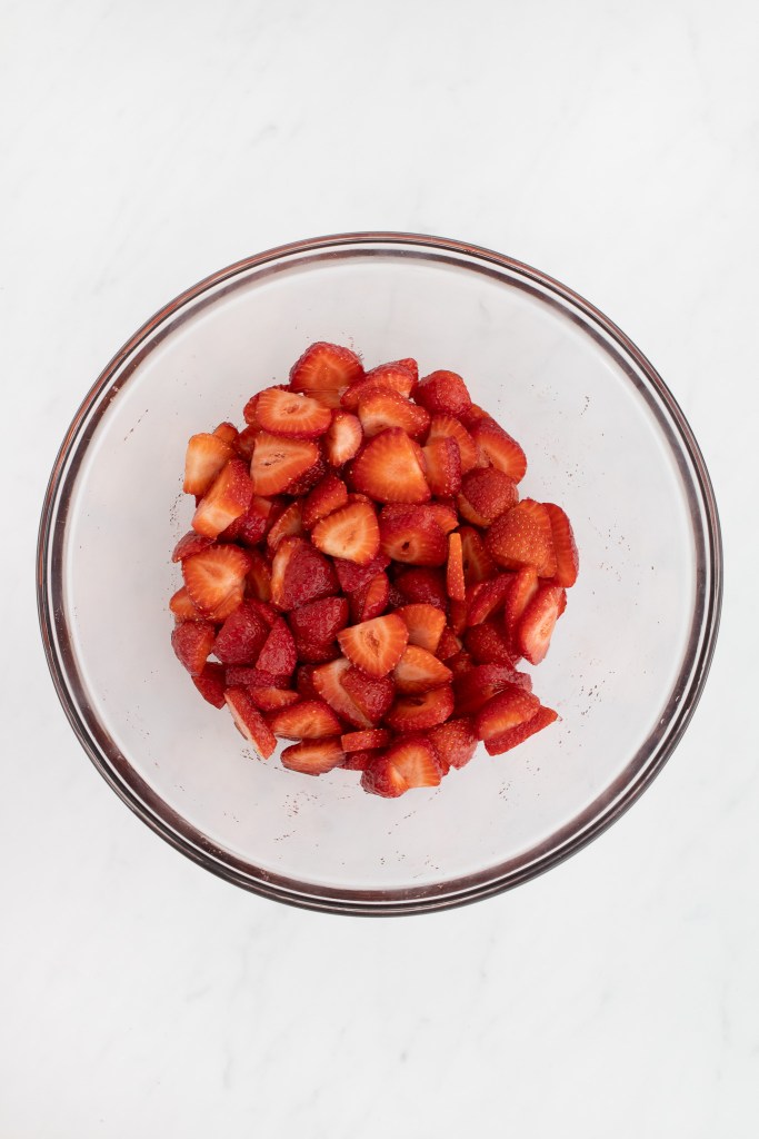 A clear glass bowl filled with sliced macerating strawberries sits on a white marble surface.