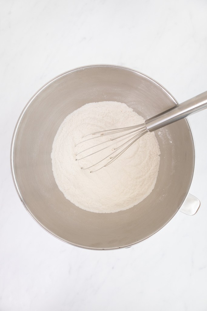 The bowl of an electric stand mixer with flour mixture inside and a metal whisk resting on top, viewed from above on a white marble surface.