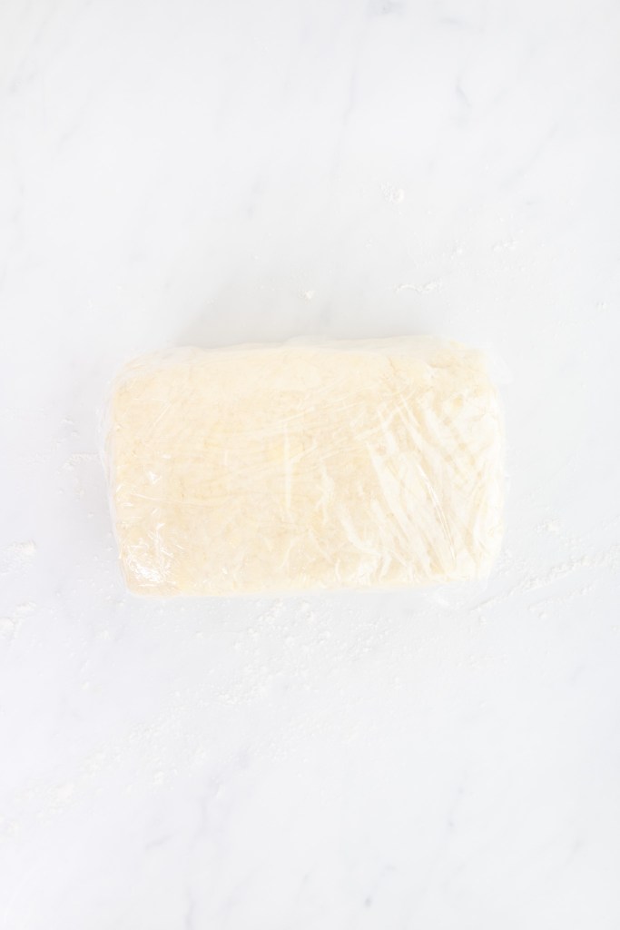 Four pieces of stacked strawberry shortcake dough wrapped in plastic wrap sits on a white marble surface.