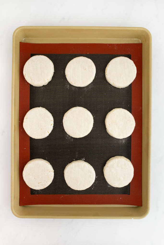 Nine unbaked round strawberry shortcakes are arranged evenly on a silicone baking mat placed on a baking sheet.