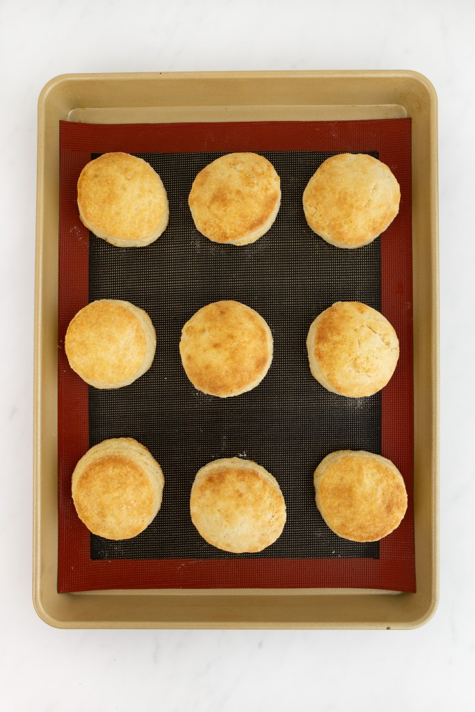 Nine golden brown strawberry shortcakes are arranged evenly in rows on a baking sheet lined with a silicone mat.