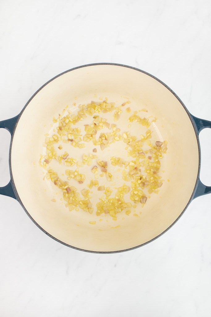 Diced shallot and garlic sautéing in oil in a large dutch oven on a white marble surface.