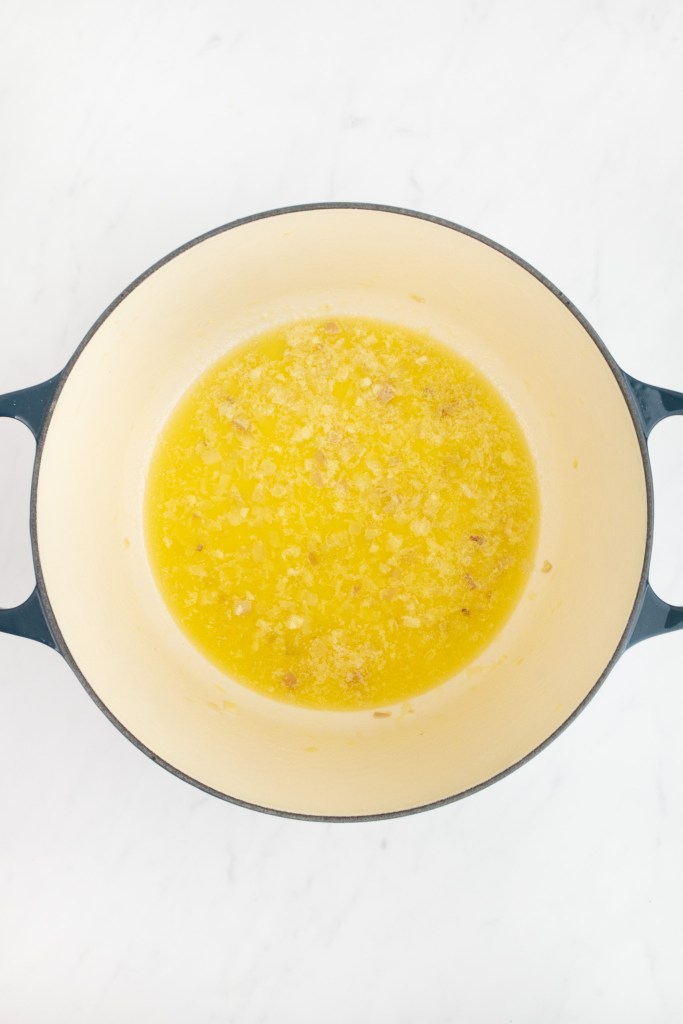 A dutch oven with melted butter and sautéed minced shallot and garlic, viewed from above, on a white marble surface.