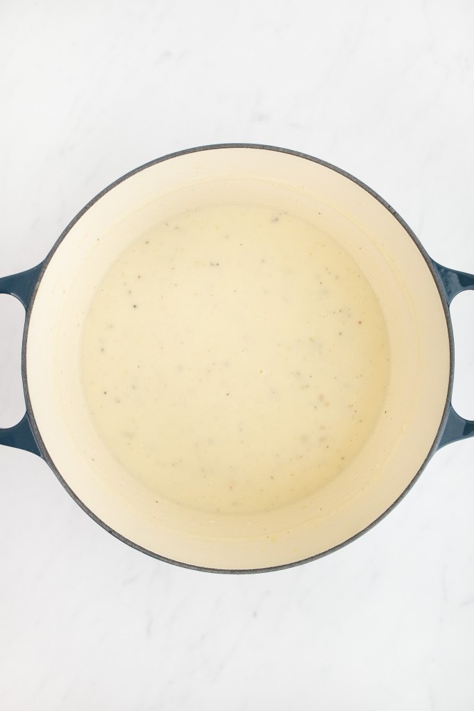 A dutch oven filled with creamy white sauce sits on a white marble surface, viewed from above.