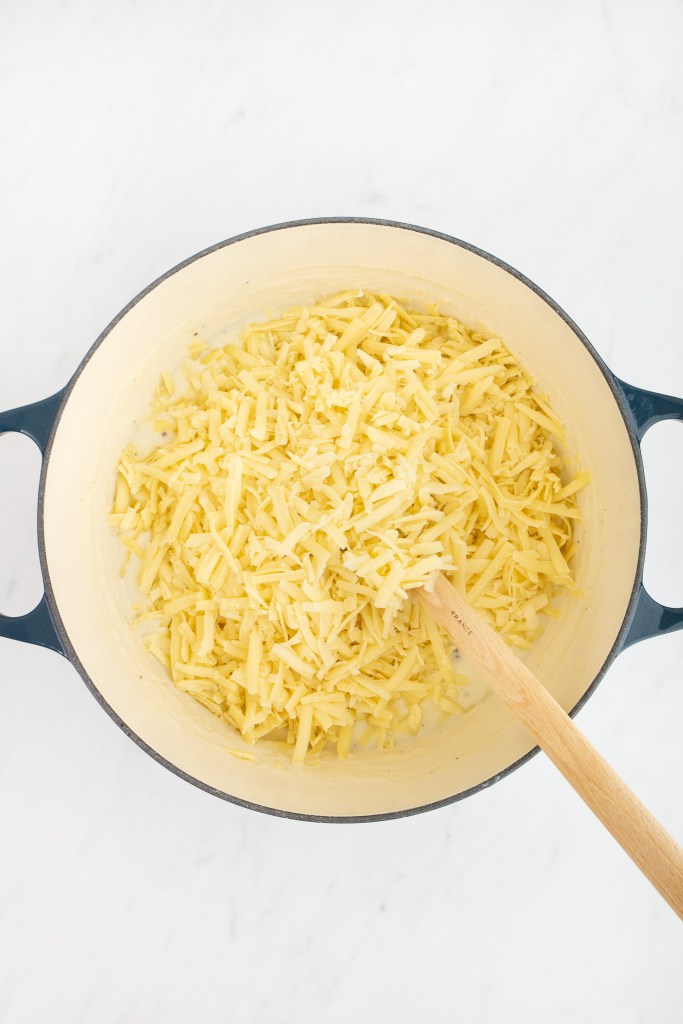 A dutch oven filled with creamy white sauce with cheese being stirred in with a wooden spoon on a white marble surface.
