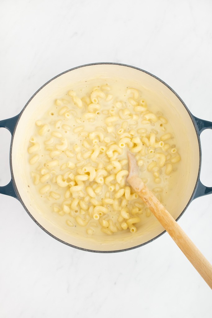 A dutch oven of macaroni and cheese with a creamy sauce, stirred with a wooden spoon, sits on a white marble surface.