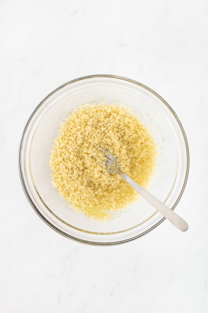 A glass bowl containing a mixture of melted butter and panko breadcrumbs on a white marble surface.