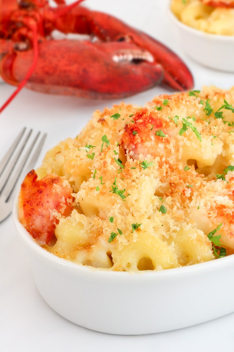 Baked lobster macaroni and cheese in a white dish, topped with breadcrumbs and chopped parsley, with a whole cooked lobster in the background.