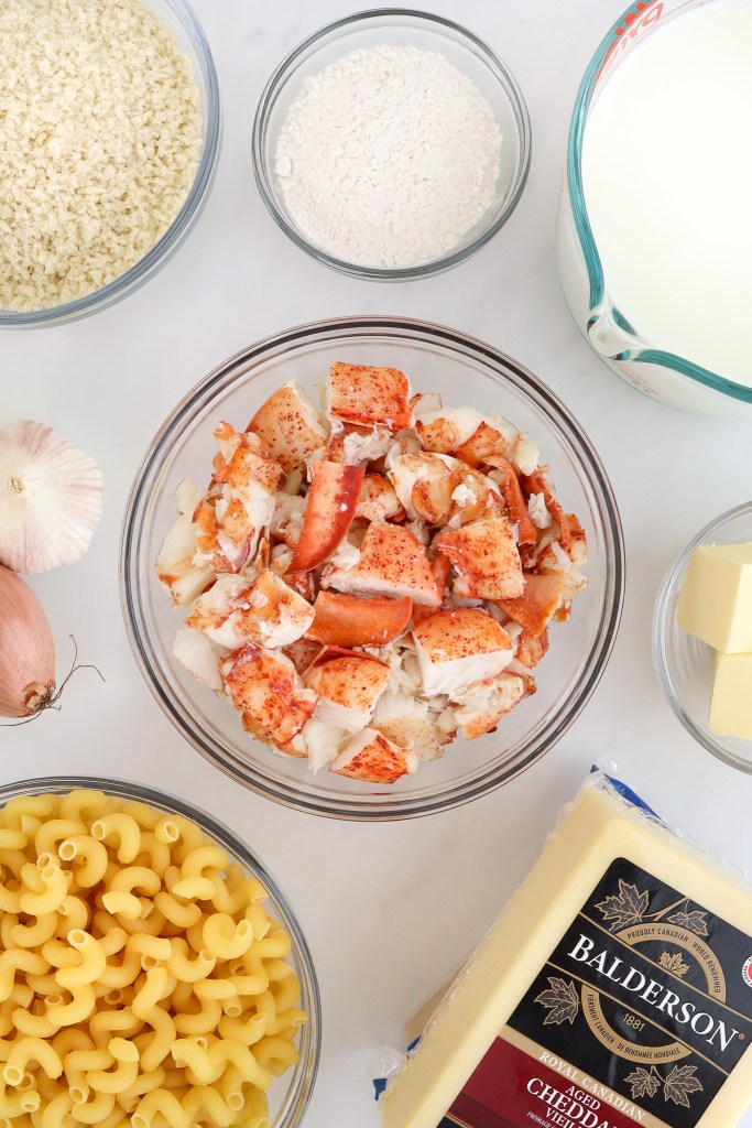 Ingredients for making lobster mac and cheese including: dry pasta, lobster meat, cheddar cheese, butter, milk, flour, panko crumbs, garlic, shallot. All items arranged on a white marble surface.