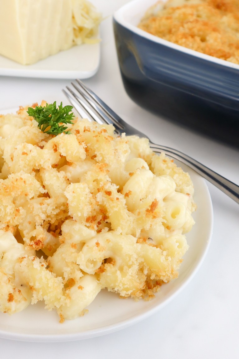 A plate of baked macaroni and cheese topped with golden breadcrumbs, garnished with parsley, with a fork and casserole dish nearby.