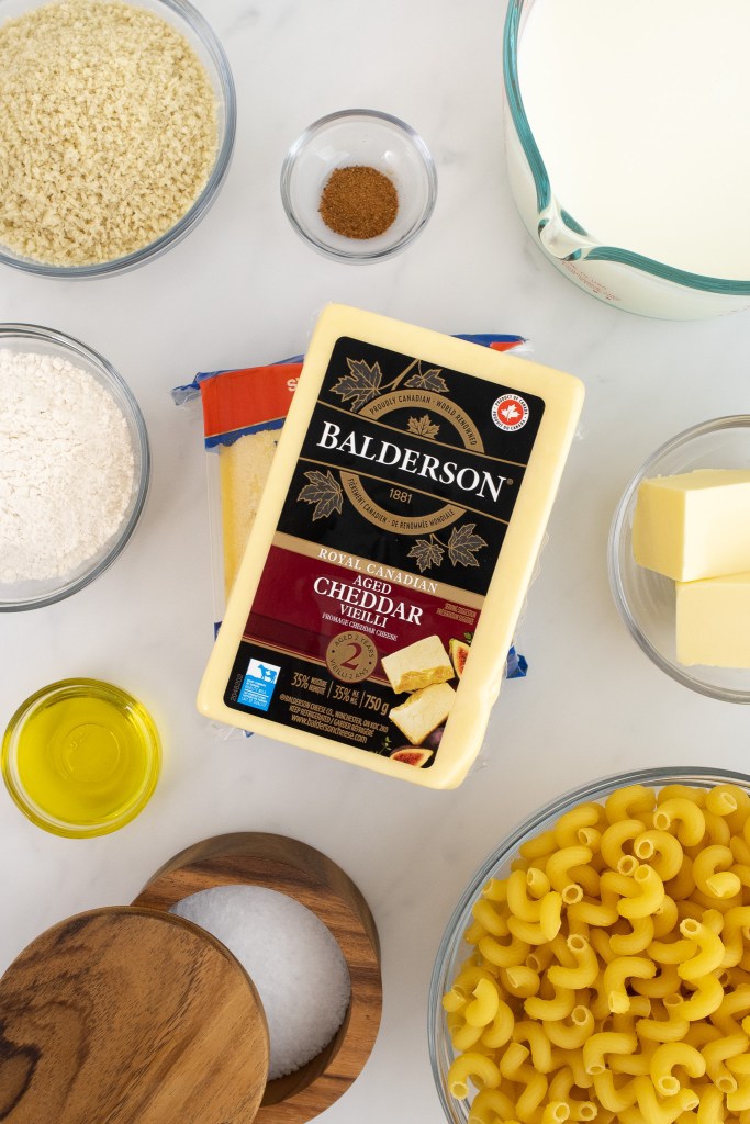 Ingredients for making Mac and cheese including: a block of Balderson cheddar, dry pasta, flour, butter, milk, oil, panko, salt, and spices in bowls arranged on a white marble surface.