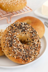 An everything bagel on a white plate with other bagels and a block of cream cheese in the background.