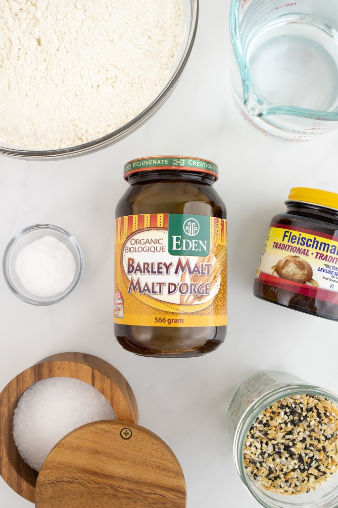 Ingredients for bread making, including flour, water, barley malt, yeast, salt, and everything bagel seasoning on a white surface.