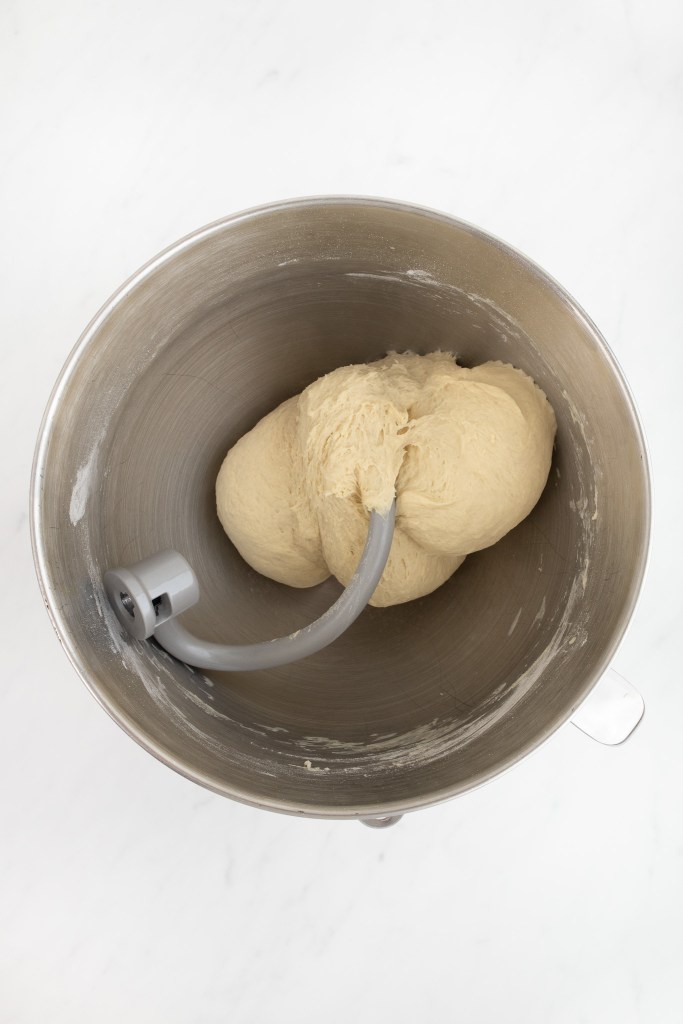 The bowl of an electric stand mixer with a ball of bagel dough being kneaded by a dough hook.