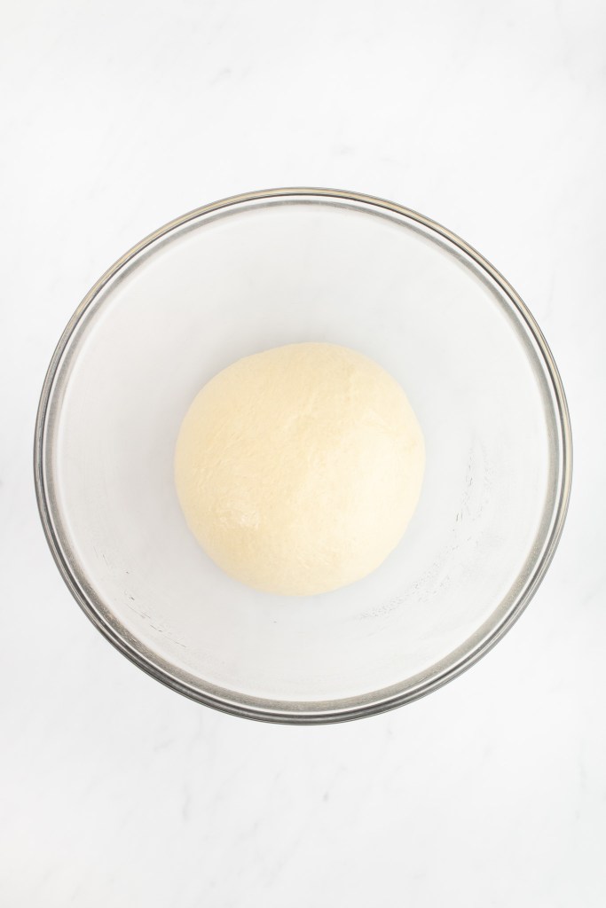 A smooth ball of unrisen bagel dough in a clear glass mixing bowl.