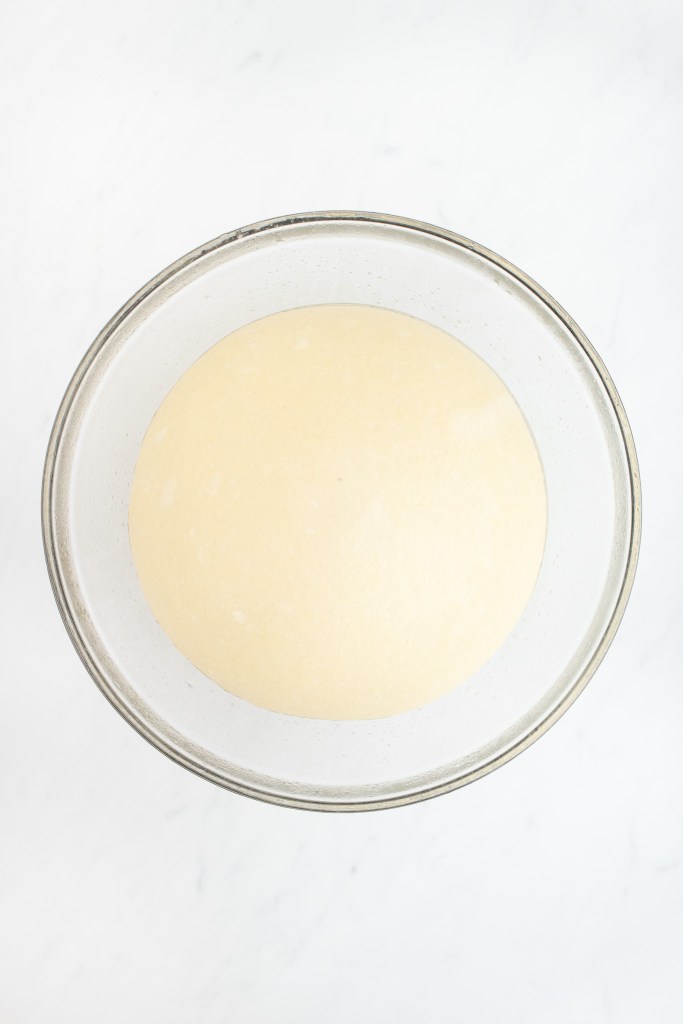 A ball of risen bagel dough in a glass bowl.