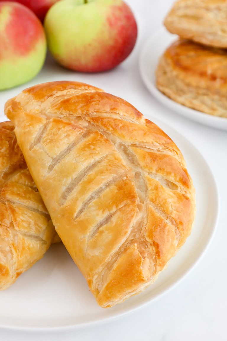 Golden-brown apple turnover on a white plate, with fresh apples and more turnovers in the background.